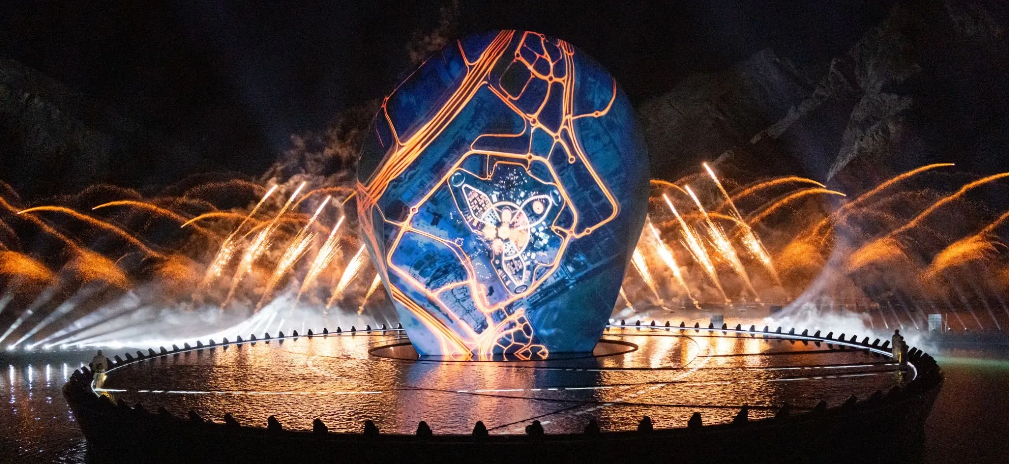 A vast egg-shaped screen standing on water, carrying a city map in blue and orange, fireworks rising behind it